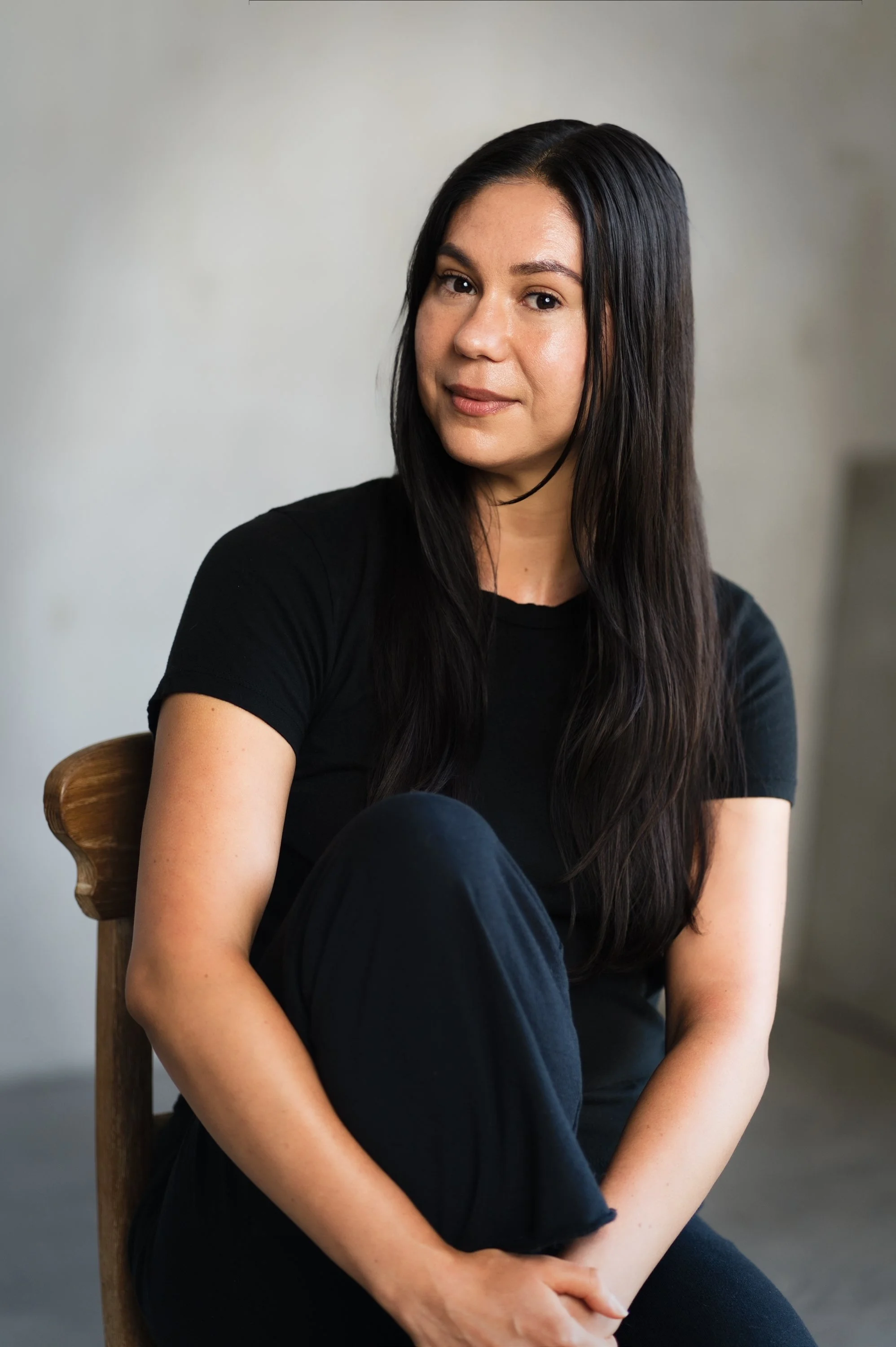 Portrait of a woman seated on a wooden chair against a neutral background.