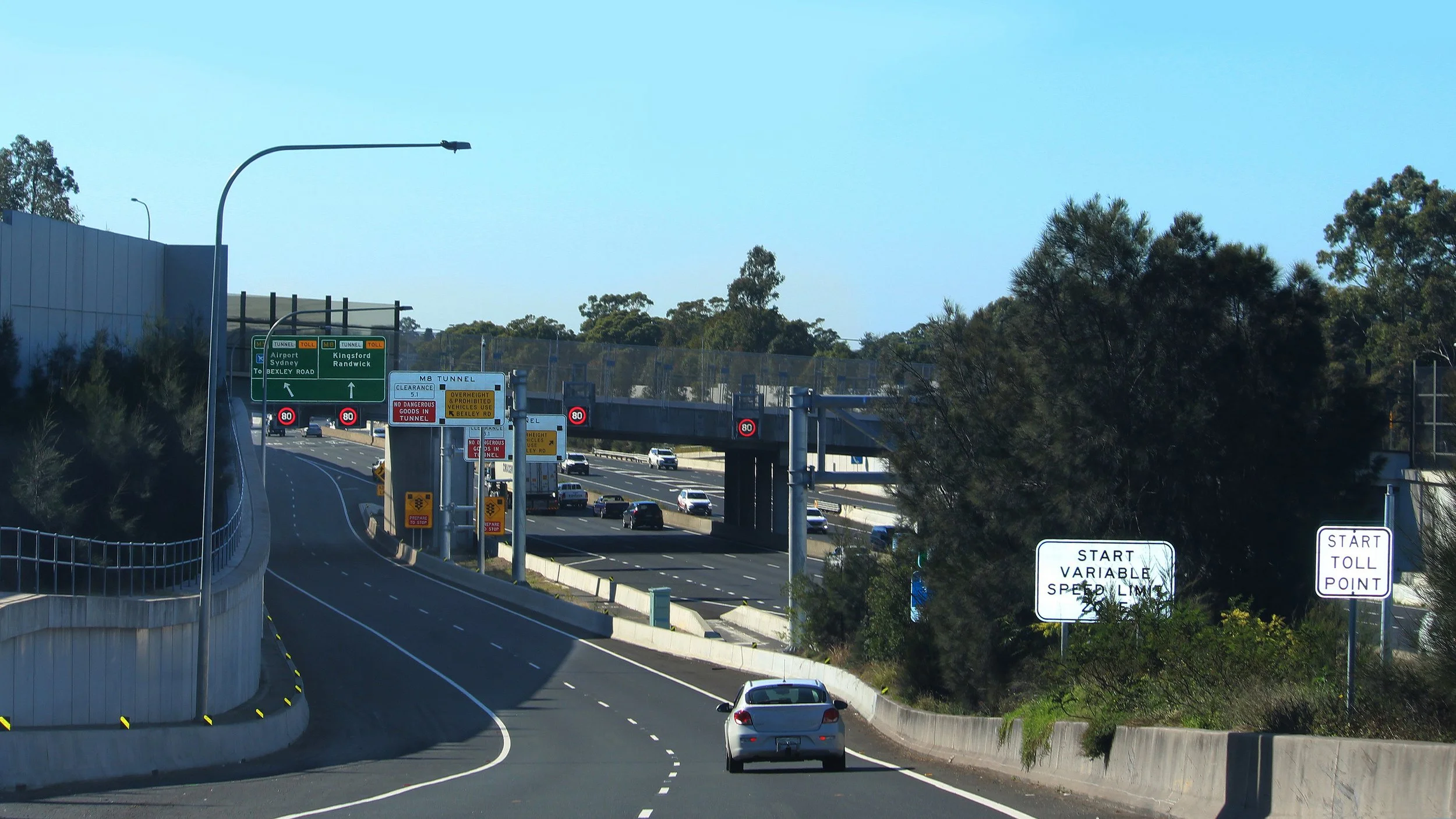 Multiple highway signs and vehicles on a highway near trees and overhead structure, with clear weather.