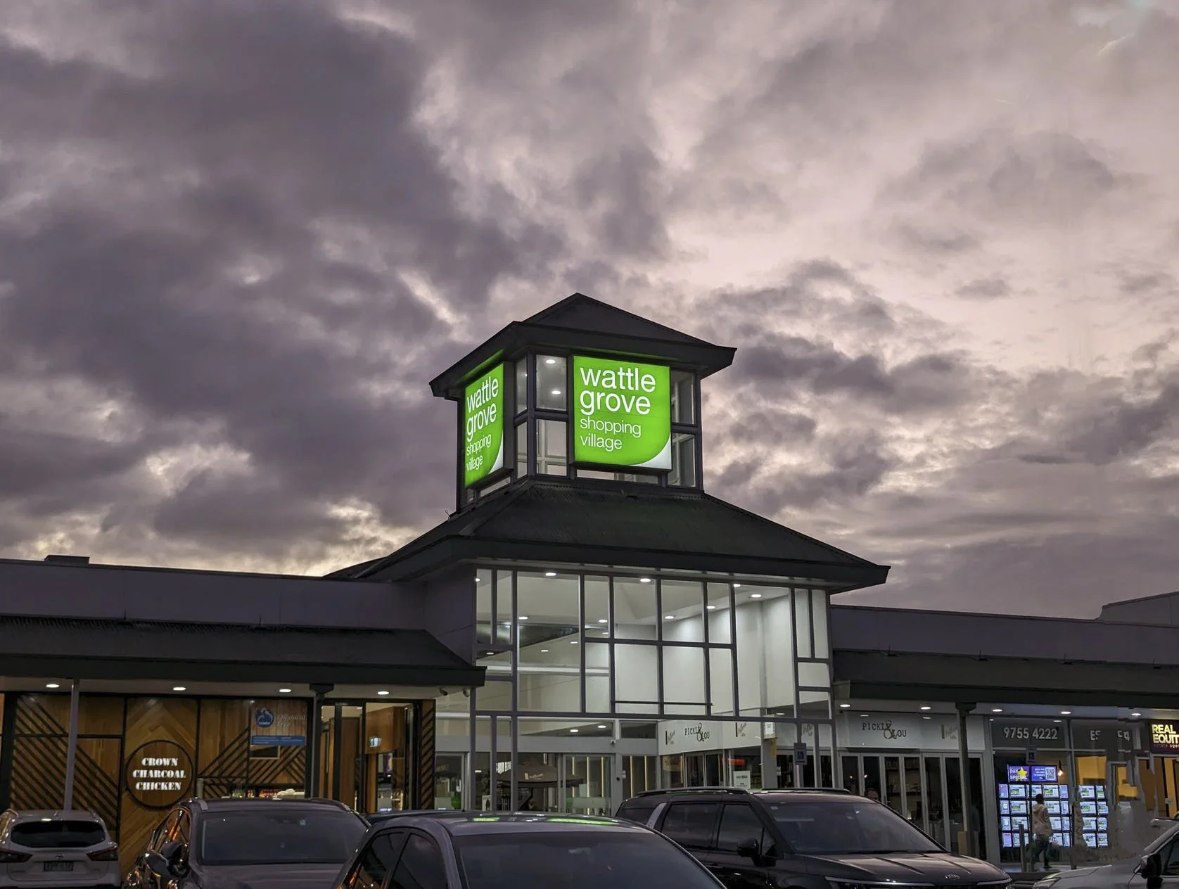 Exterior view of Wattle Grove Shopping Village at dusk with a modern glass-front building and a sky filled with dark clouds, featuring a prominent green sign displaying the shopping center's name.