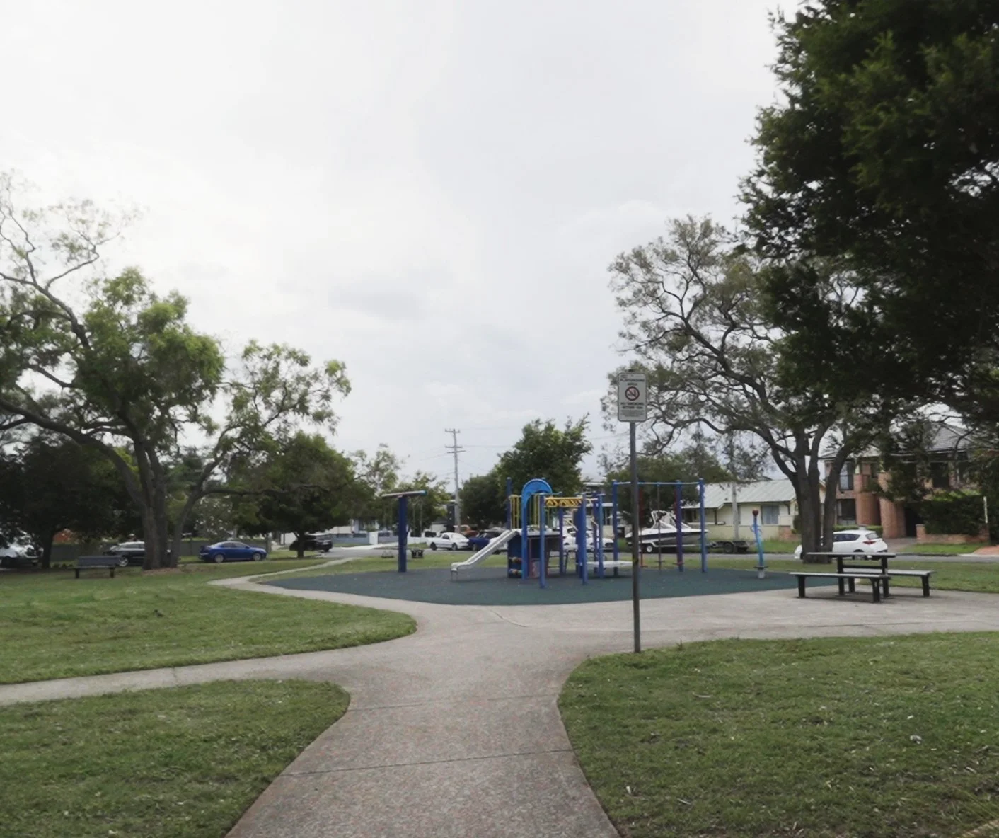 A playground in a park with a winding concrete pathway leading to it. The playground has blue and yellow equipment, and there are trees and houses in the background. A sign indicates no smoking or pets allowed.