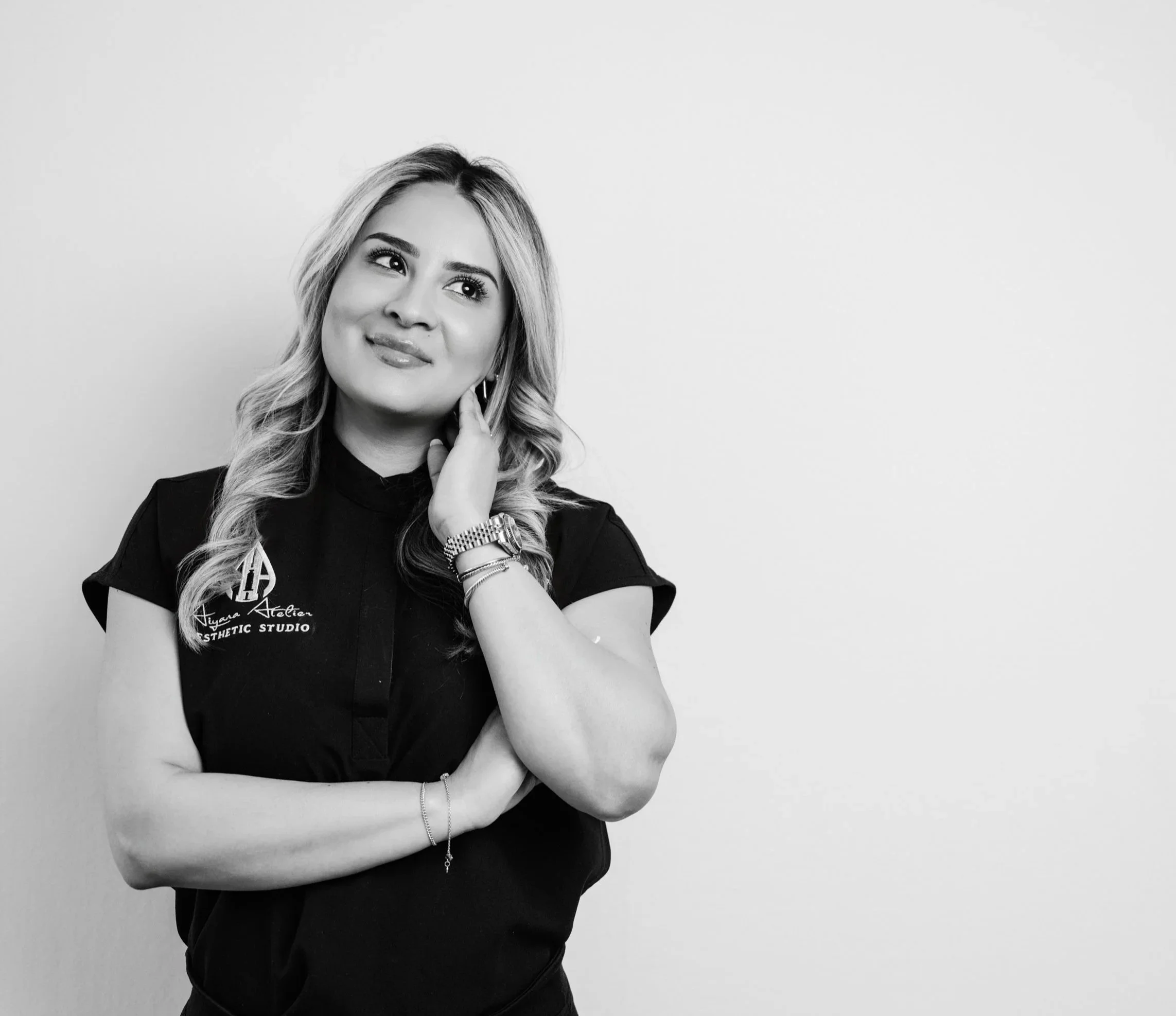 Black and white portrait of a woman with long wavy hair, wearing a black shirt with text, accessories, and a watch, against a plain light background.