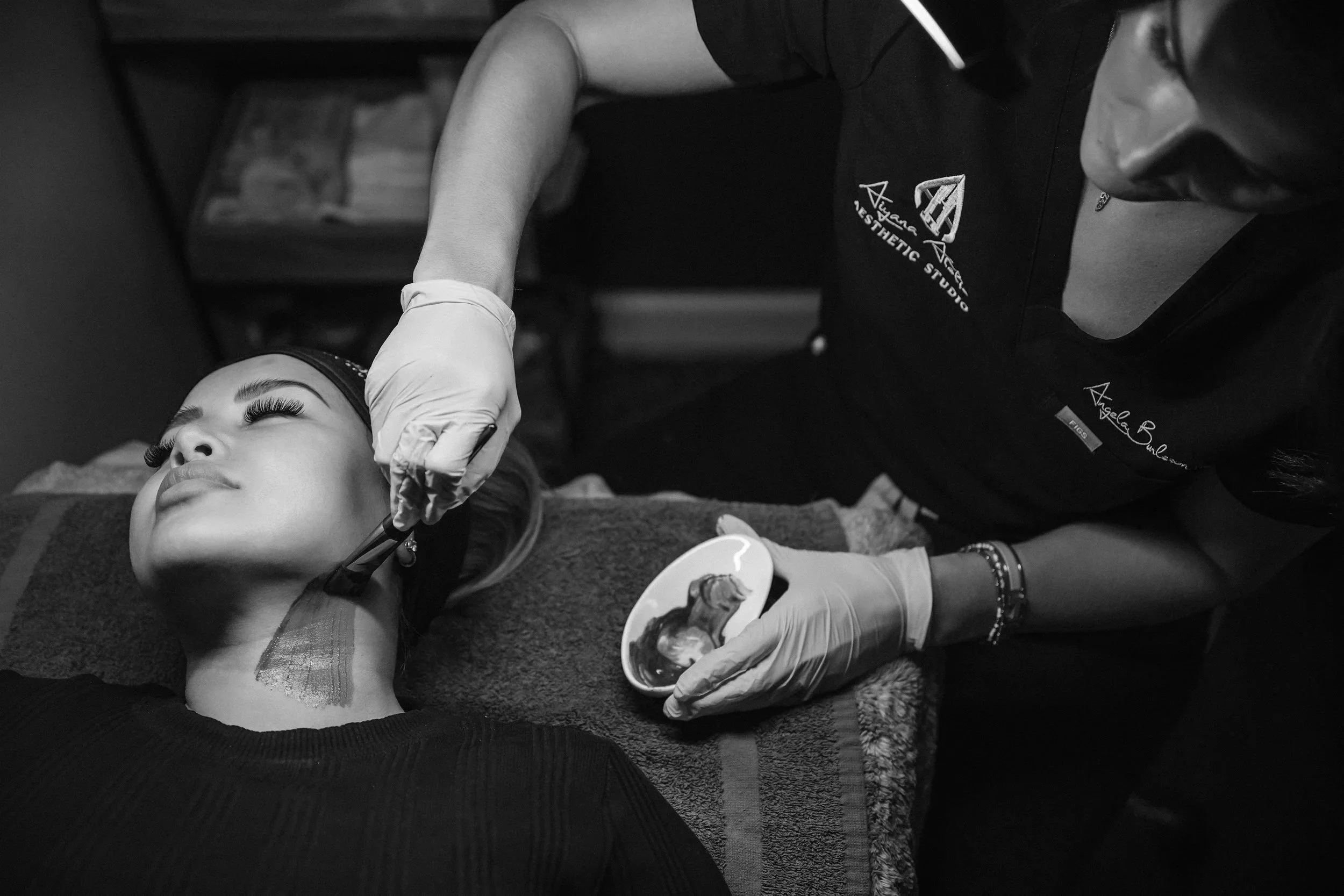 A woman lying down receiving a cosmetic or medical procedure on her face from an aesthetic technician or doctor, who is wearing gloves and a uniform, in a clinical setting.