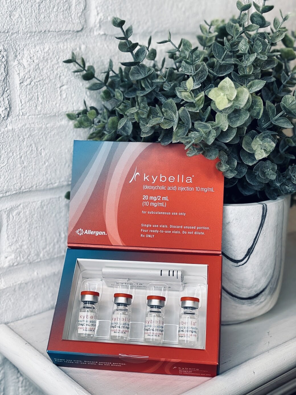 A box of Kybella, an injectable treatment for submental fat reduction, containing four vials with red caps, displayed next to a potted green plant against a white brick wall.