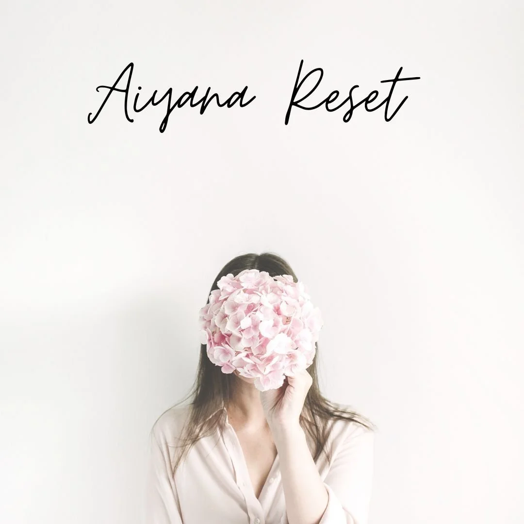 A woman holds a pink flower in front of her face, obscuring her facial features, with the words 'Aiyana Reset' written in black script at the top of the image.