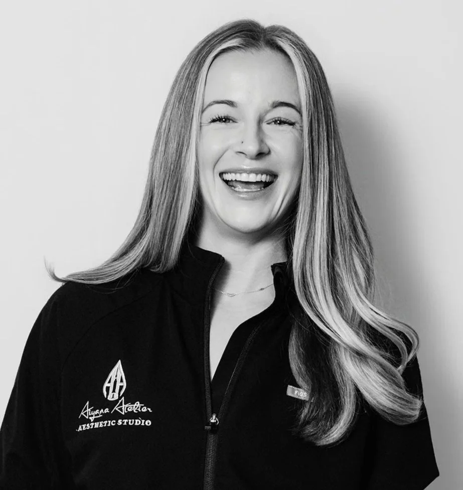 A woman smiling and winking, with long blonde hair, wearing a black zip-up jacket with the logo and text for 'Ayana Aesthetic Studio' on the chest.