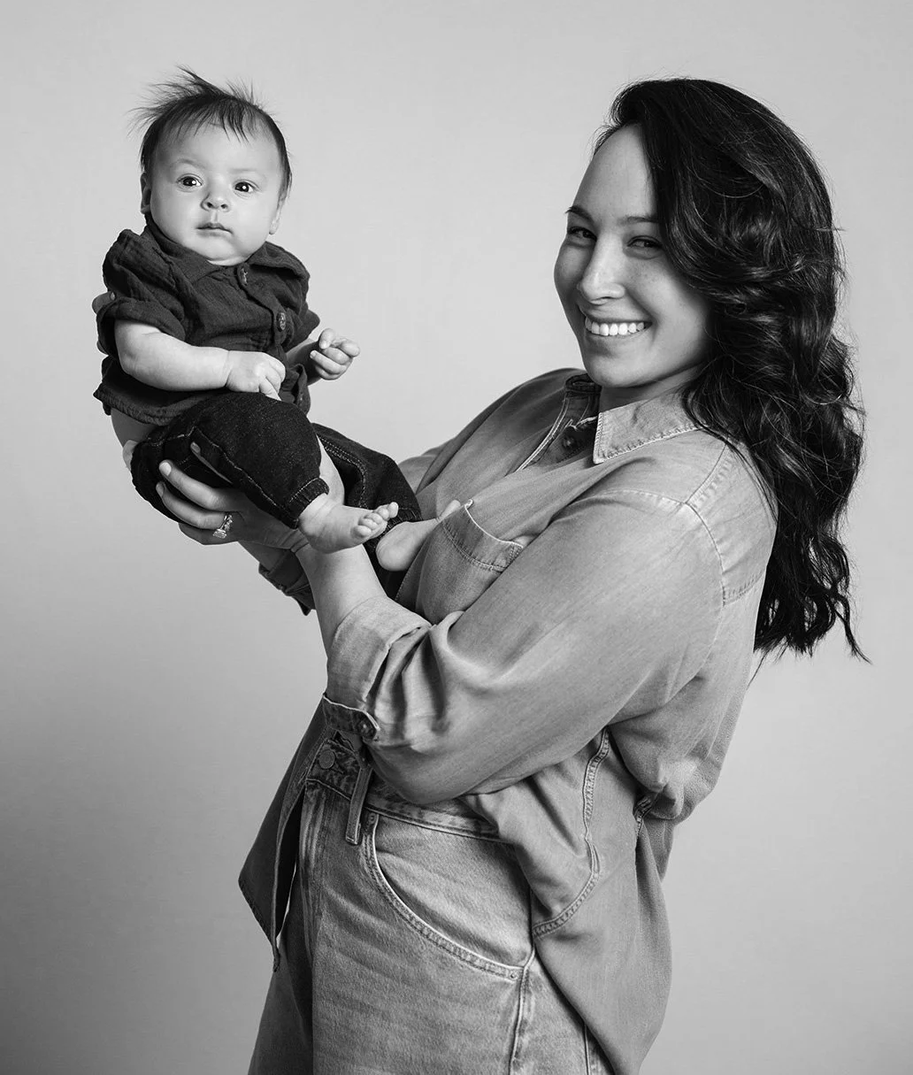 A woman with long curly hair smiling while holding a small baby in her arms in front of a plain background.