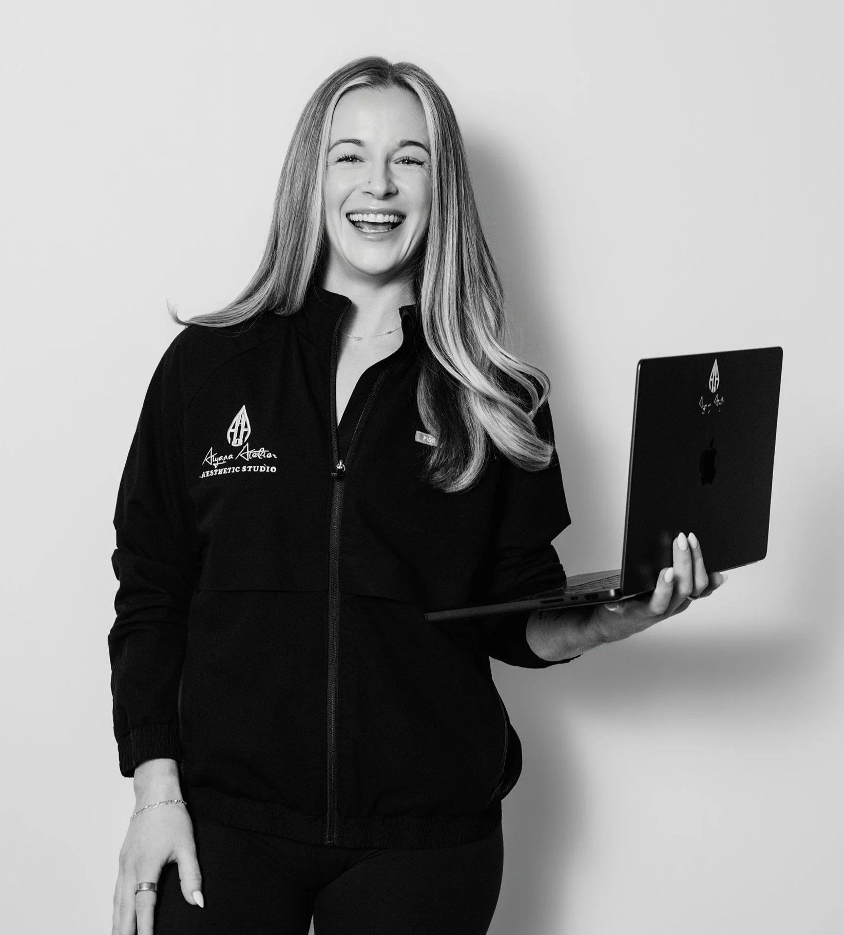 Black and white photo of a woman smiling and holding a laptop, wearing a black zip-up jacket with the logo and text 'Alyana Athletic Aesthetic Studio' on it.
