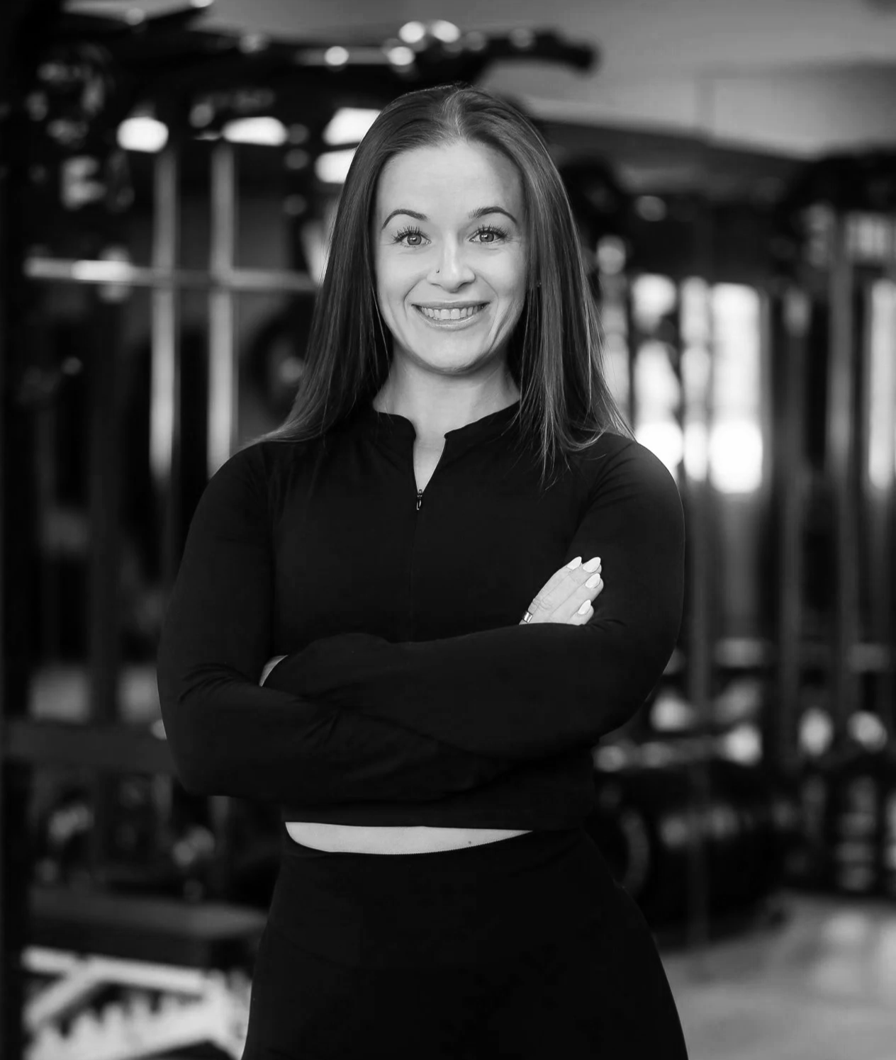 A woman with long straight hair, smiling, standing with arms crossed in a gym with workout equipment in the background.
