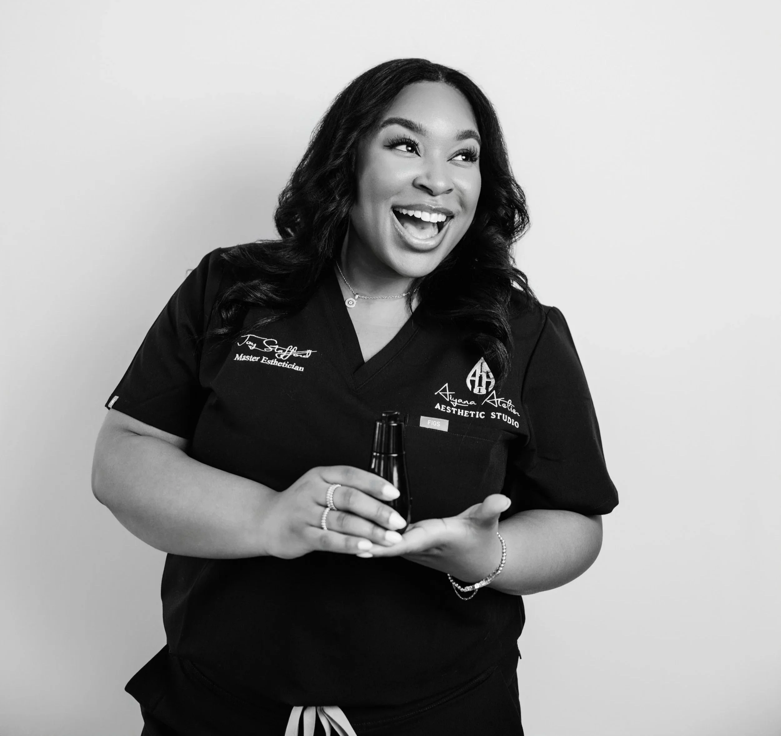 Black and white photo of a smiling woman with long curly hair wearing a black medical scrub top with embroidered text, jewelry including rings and bracelets, holding a small bottle, against a plain light background.