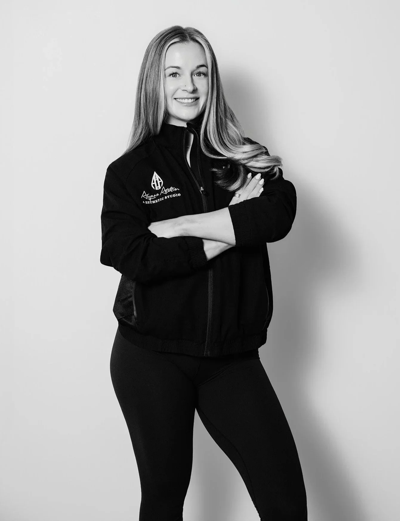 Black and white portrait of a young woman with long hair, smiling, wearing a workout jacket with a logo on the chest and black leggings, standing with arms crossed against a plain background.