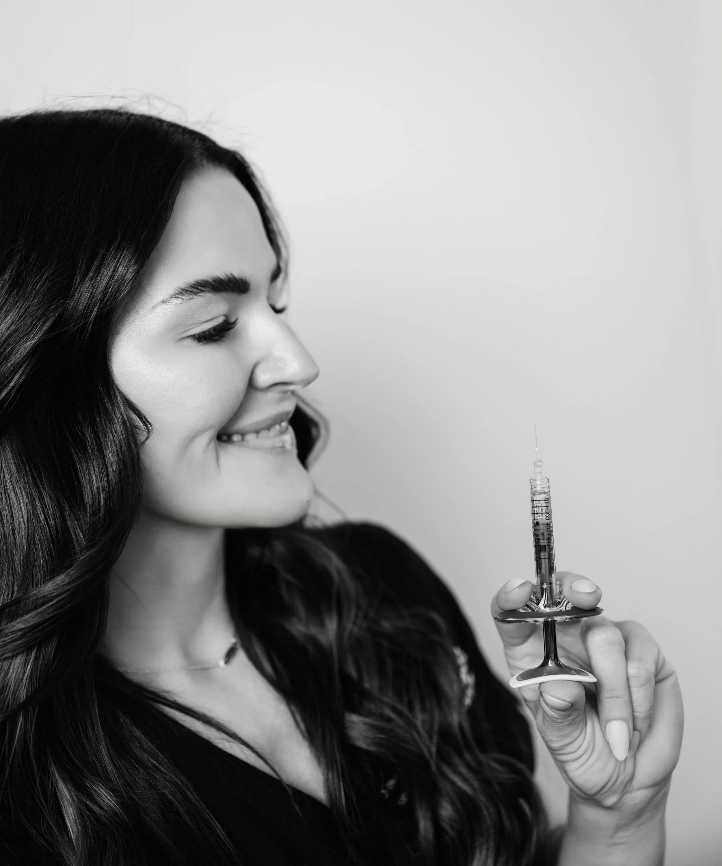 A woman with dark hair smiling while holding a syringe with a needle, in black and white.