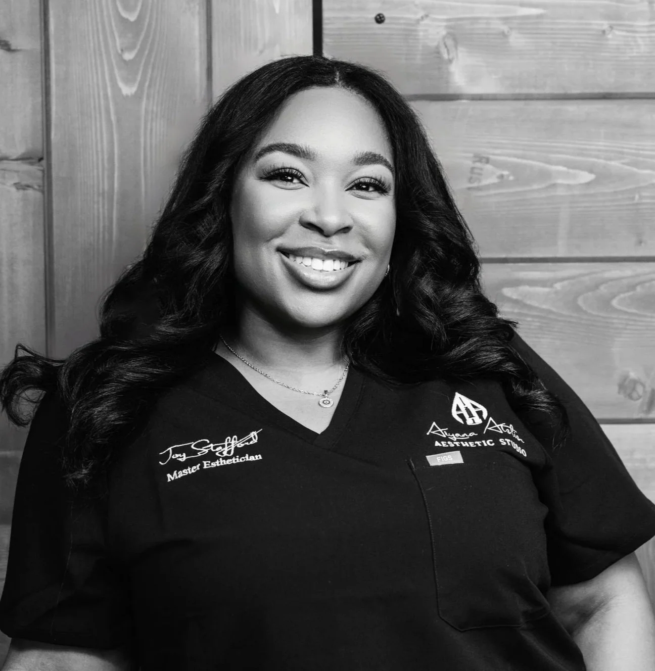 Portrait of a woman smiling in a professional uniform with embroidered text, against a wooden background in black and white.