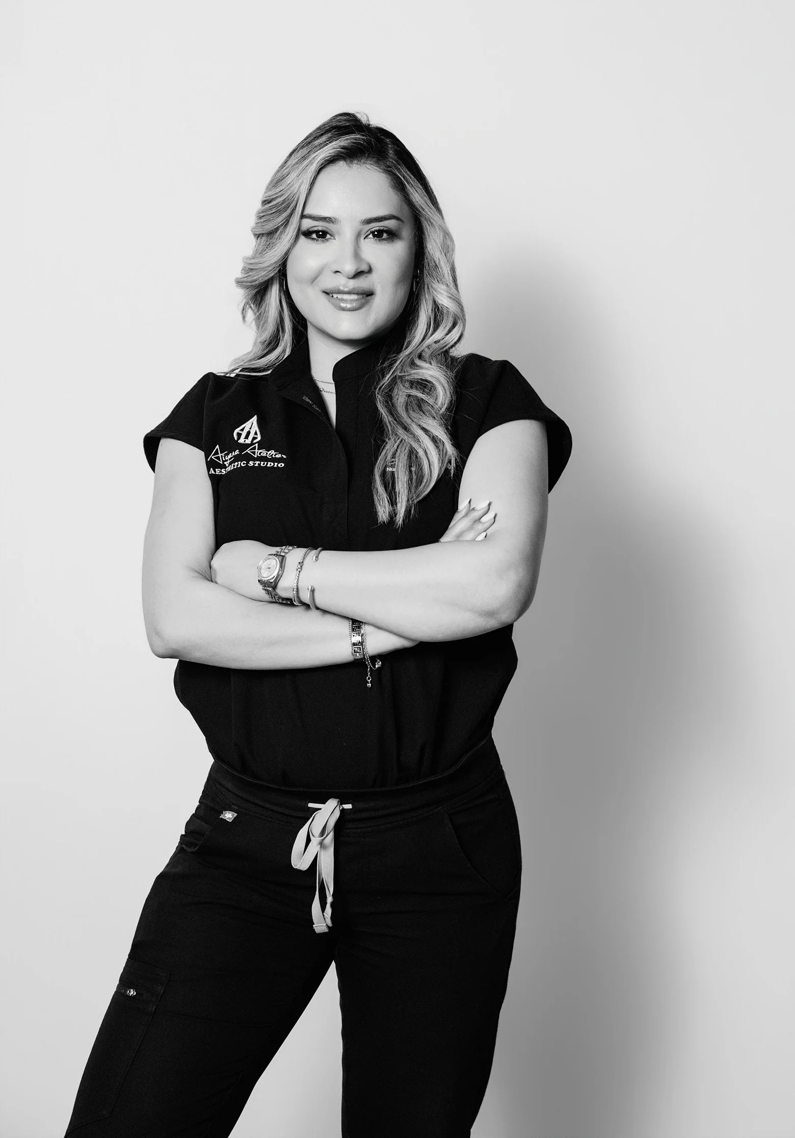 Black and white photo of a woman standing with arms crossed, wearing a scrubs top with a logo and text, and black pants with a drawstring, smiling slightly, against a plain background.