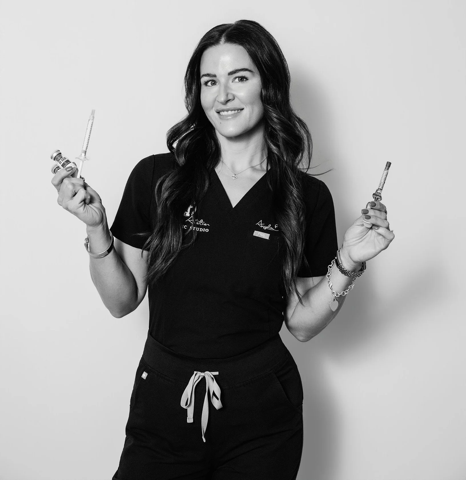 A woman with long dark hair, wearing a black T-shirt with embroidery, black drawstring pants, holding two syringes, smiling, against a plain background.