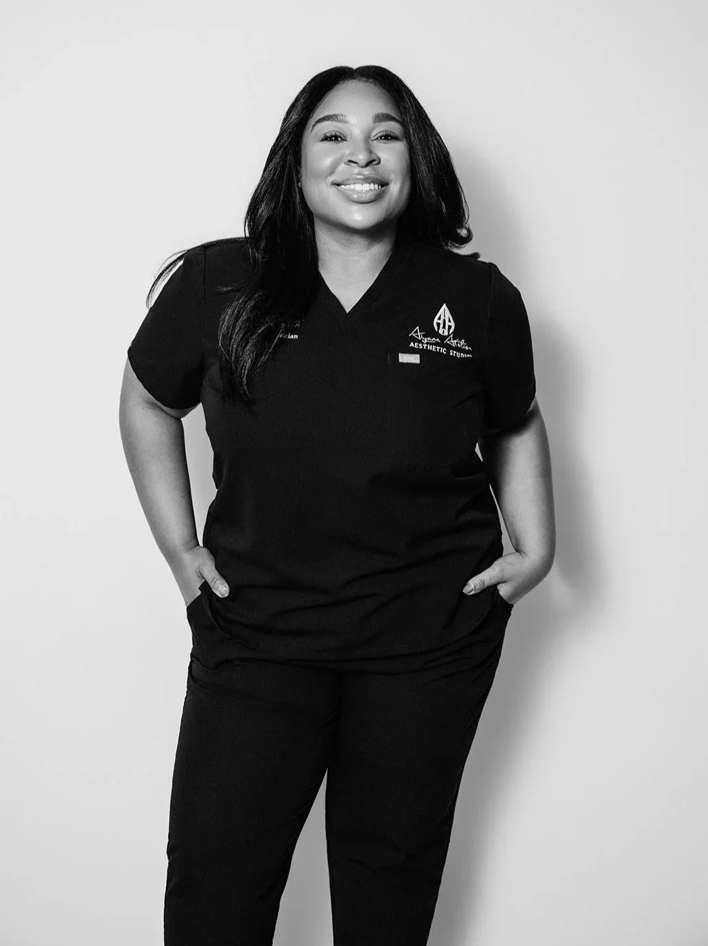 A woman smiling in black medical scrubs with hands in pockets, standing against a plain background.