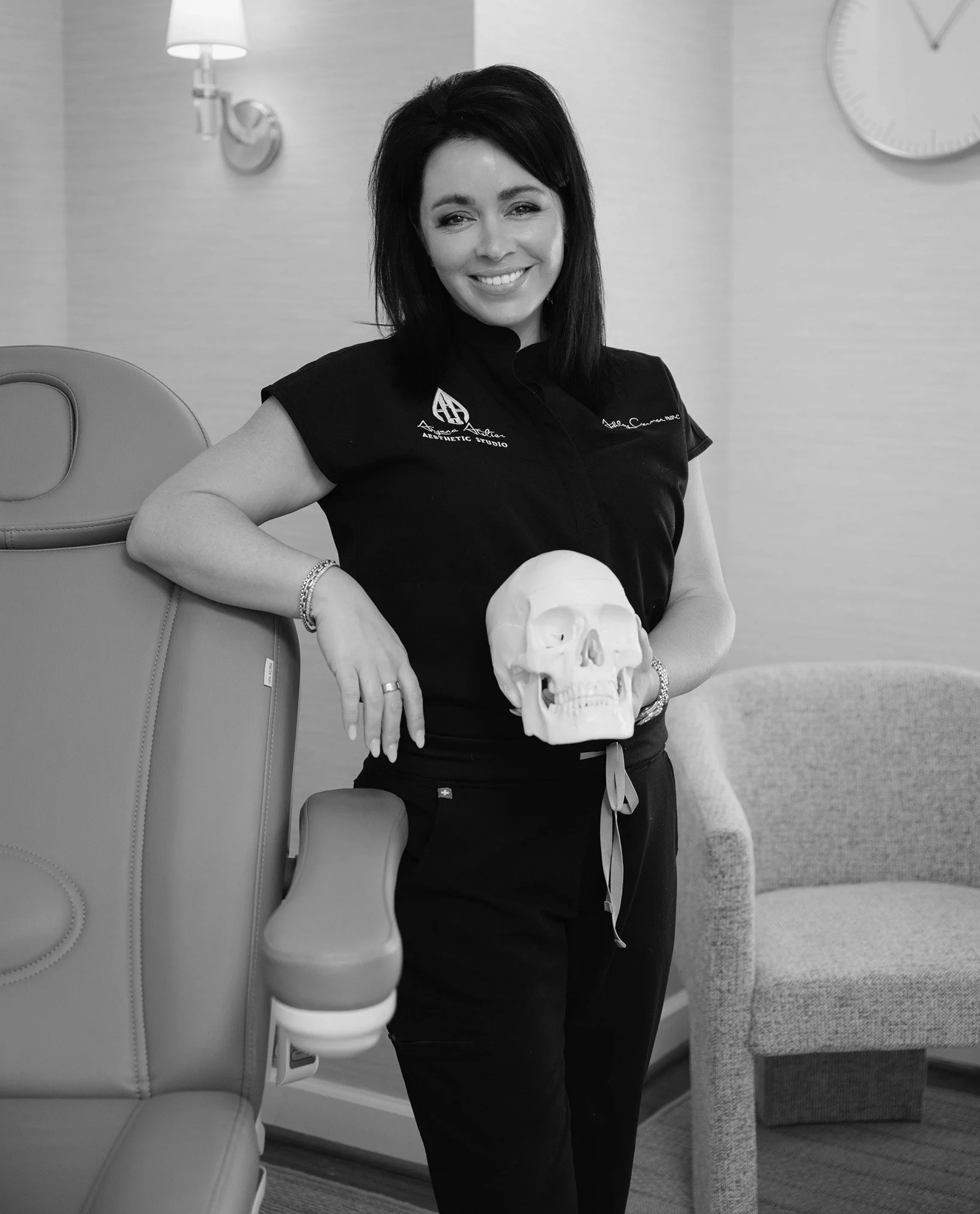 A woman with dark hair is sitting in a room, smiling, holding a human skull. She is wearing a black shirt with embroidered text and a logo. The room features a light-colored wall, a wall-mounted lamp, a large clock, and a chair with a distinctive headrest.