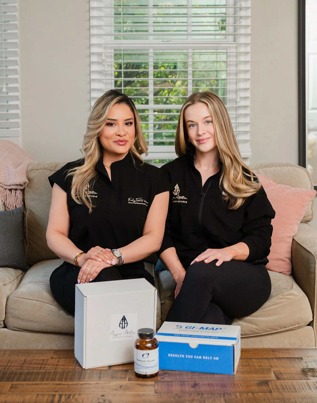 Two women sitting on a beige couch in a living room, smiling at the camera. They are wearing black shirts with logos, and there are boxes and a small bottle on the table in front of them. Behind them are large windows with blinds and greenery outside.