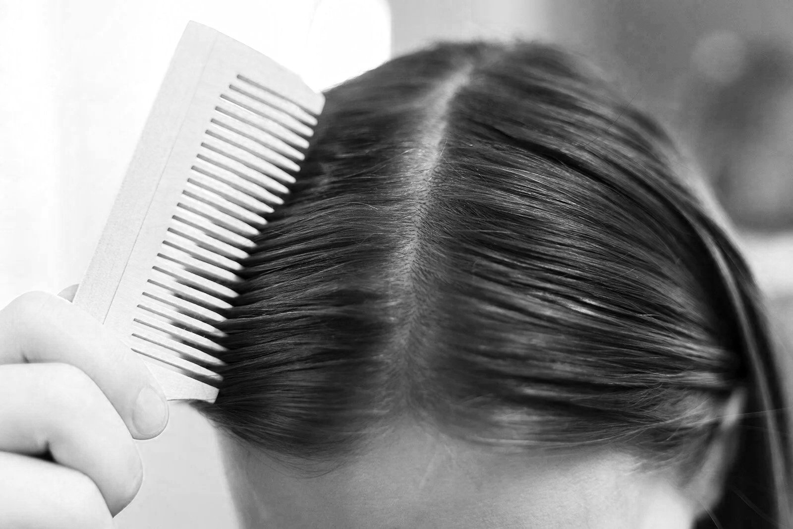 Black and white photo of a person's hair being combed with a wide-tooth comb, focusing on the top of the head and parting.