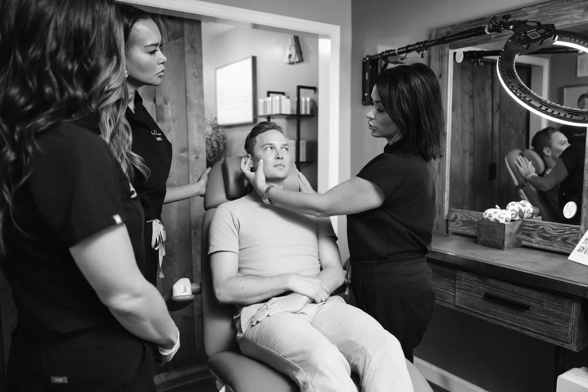 A person receiving a facial treatment from a staff member in a spa or wellness center, with other staff attending to someone else in the background.