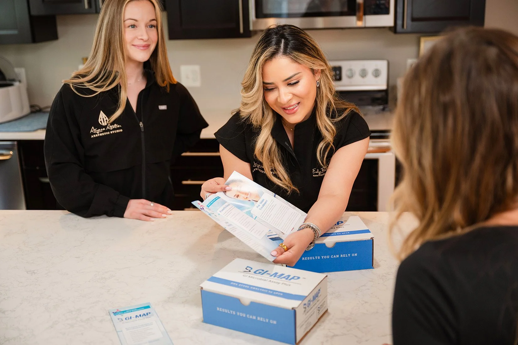 Three women, dressed in black professional attire, inside a kitchen, preparing or discussing medical test kits labeled GI-MAP, with one woman handing a test kit to another, while the third woman looks on.