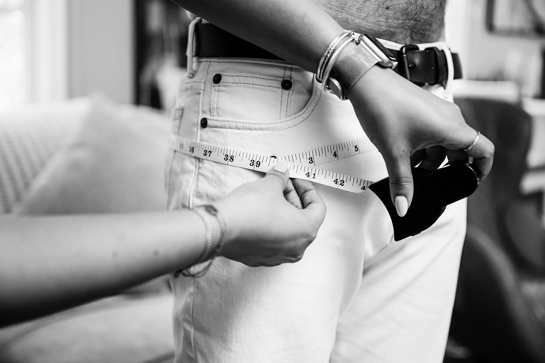 A person is measuring another person's waist with a measuring tape. The person being measured is wearing white jeans and a belt, while the measurer has a watch and a ring and is holding a smartphone in one hand.