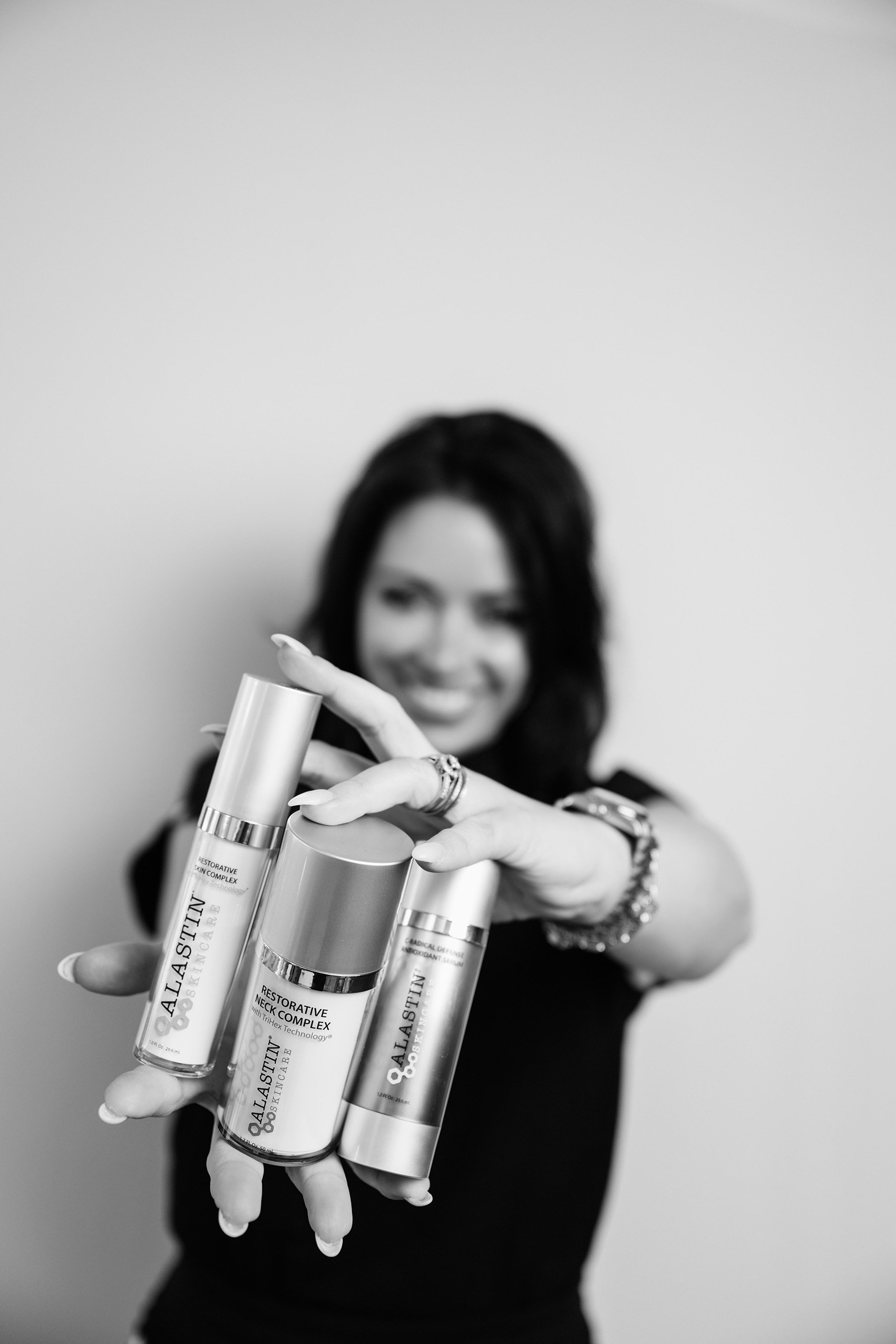 A woman with dark hair holding and displaying three bottles of skincare products in front of her. She is smiling and wearing jewelry.