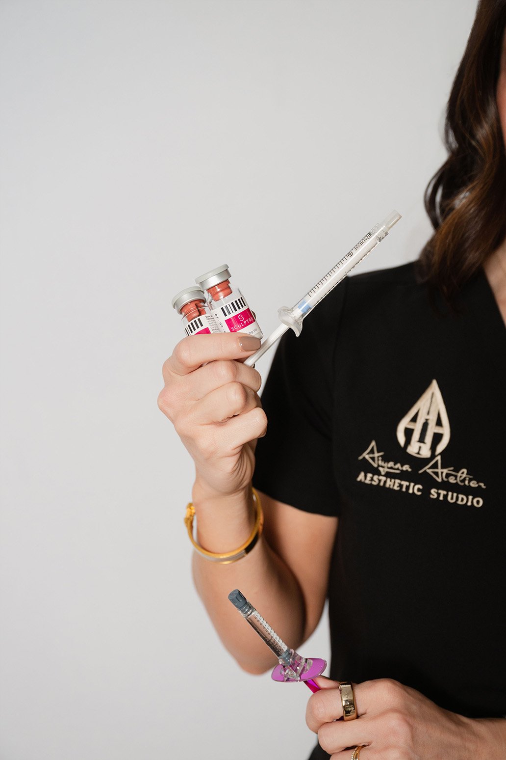 A woman holding two vials of aesthetic treatment, a syringe, and a smaller needle with a pink cap, in front of a plain gray background, wearing a black shirt with the logo of 'Aqua Atelier Aesthetic Studio'.