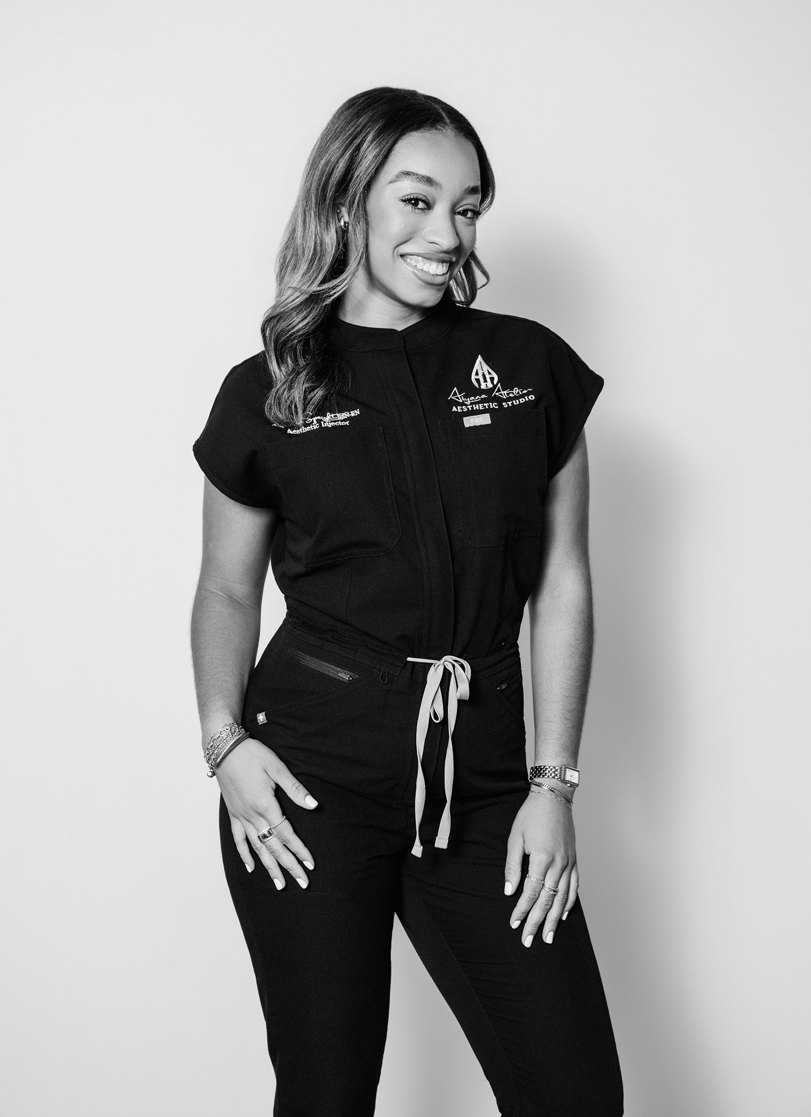 Black and white photo of a woman smiling, wearing a color-tinted shirt with embroidered patches, black pants, and jewelry, posing against a plain background.