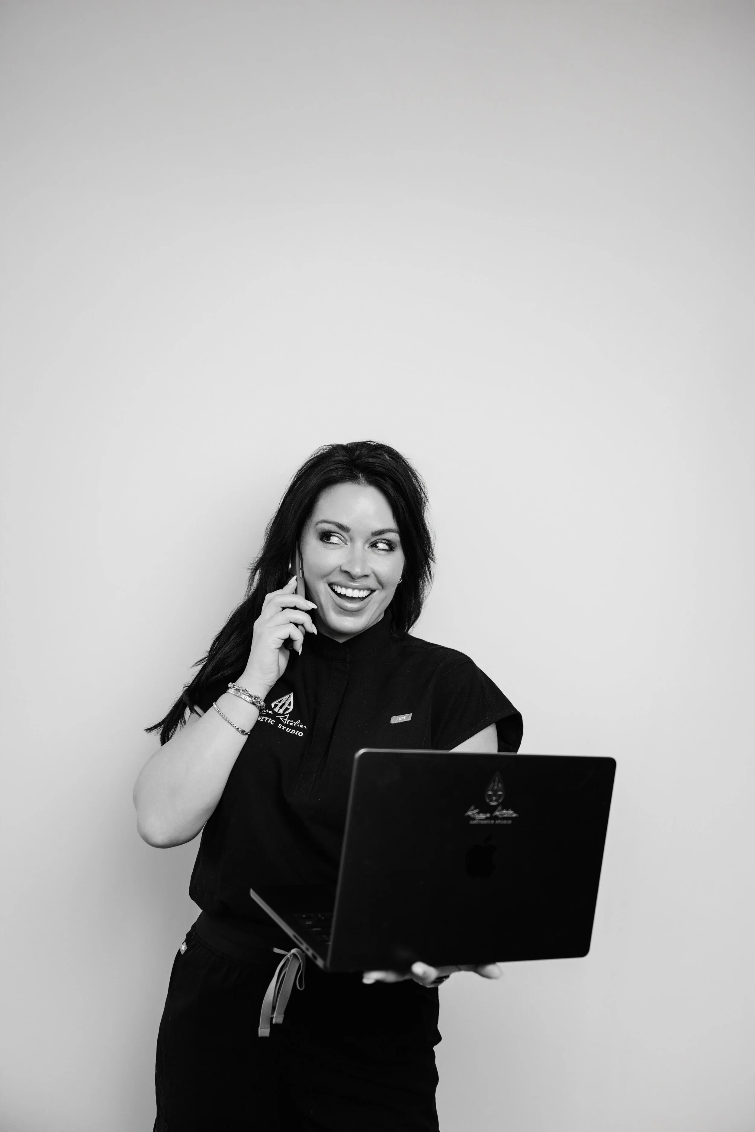 Black and white photo of a woman wearing a black medical uniform, smiling, talking on a cell phone, standing in front of a plain wall, with a laptop in front of her.