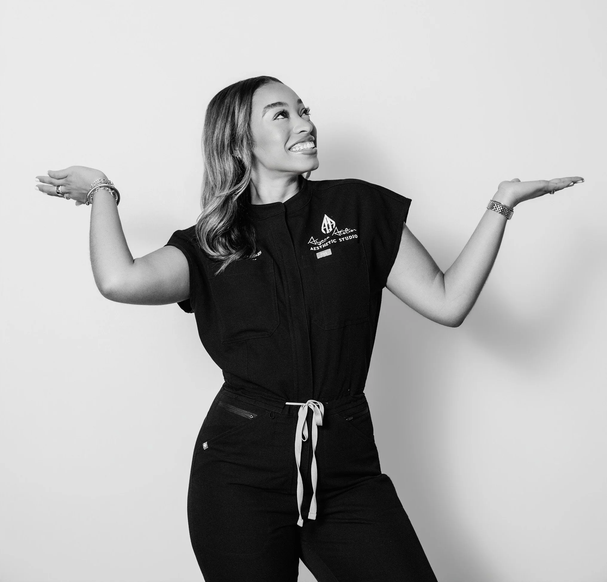 A woman wearing a black t-shirt and black pants with a white drawstring, standing against a plain light background. She is smiling and looking upward with her arms raised and hands open, wearing multiple bracelets and rings.