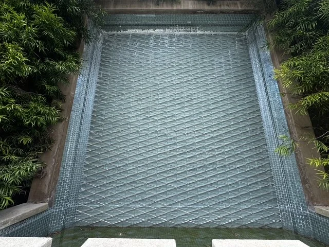 Empty swimming pool with a tiled bottom, surrounded by green bushes and trees.