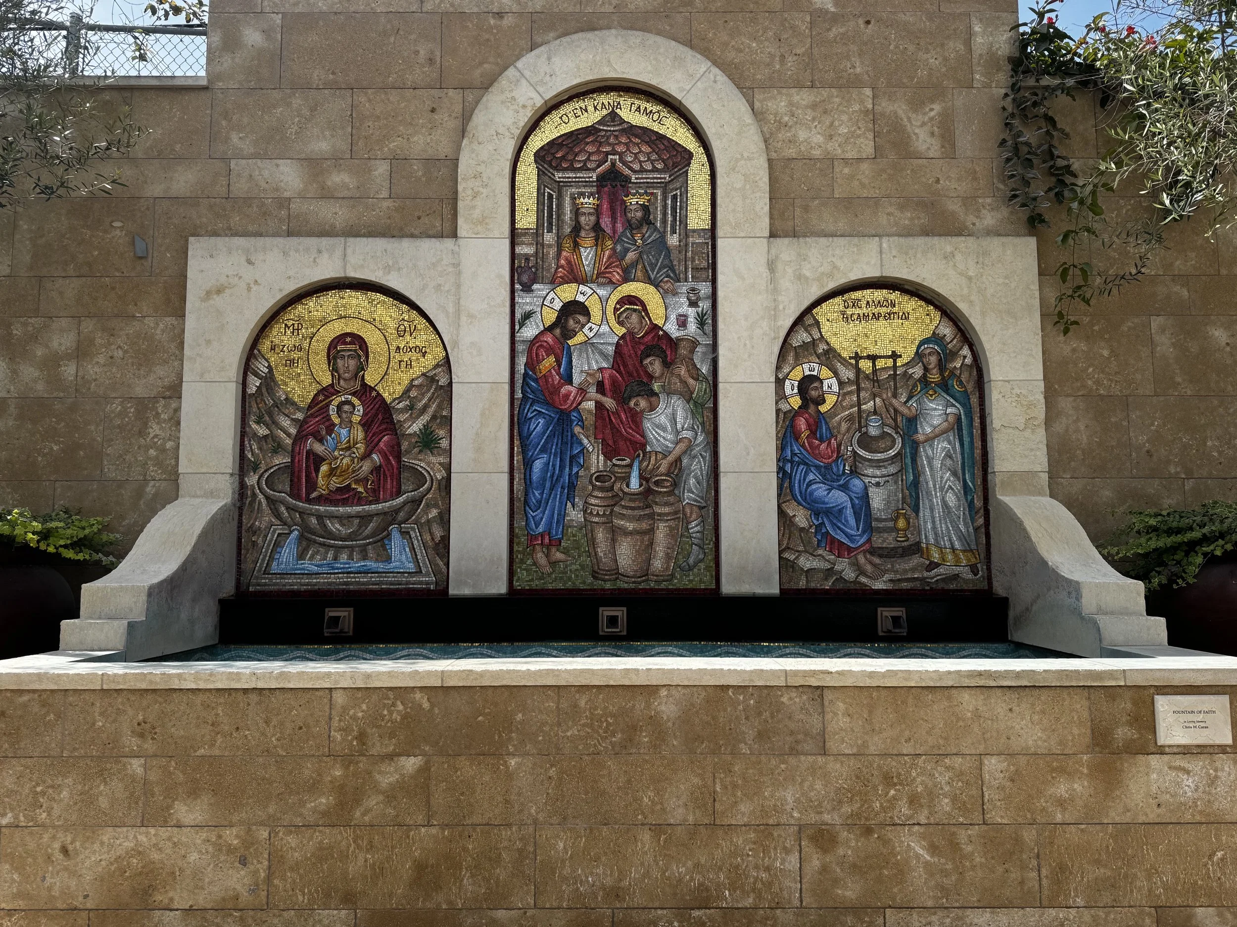 Mosaic artwork depicting religious figures, with a central scene of Jesus and a woman, and surrounding figures, set in a three-part design on a stone wall beneath an arch, with green plants on either side.