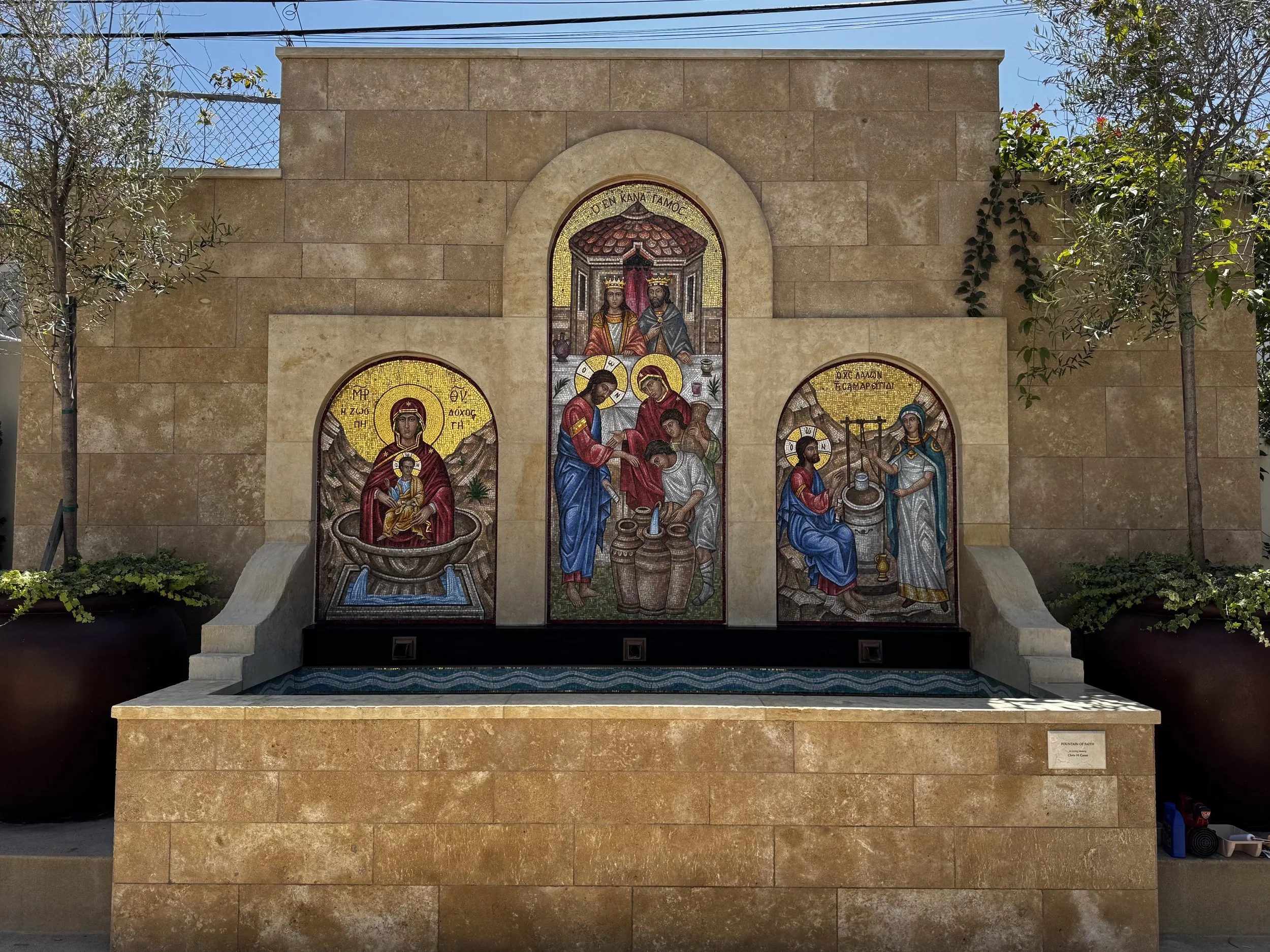 A stone fountain with religious mosaics depicting saints and biblical scenes on a beige stone wall, surrounded by trees and a clear blue sky.