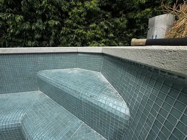 Empty swimming pool with green-tiled interior and a water jet feature, surrounded by a concrete deck and lush greenery in the background.