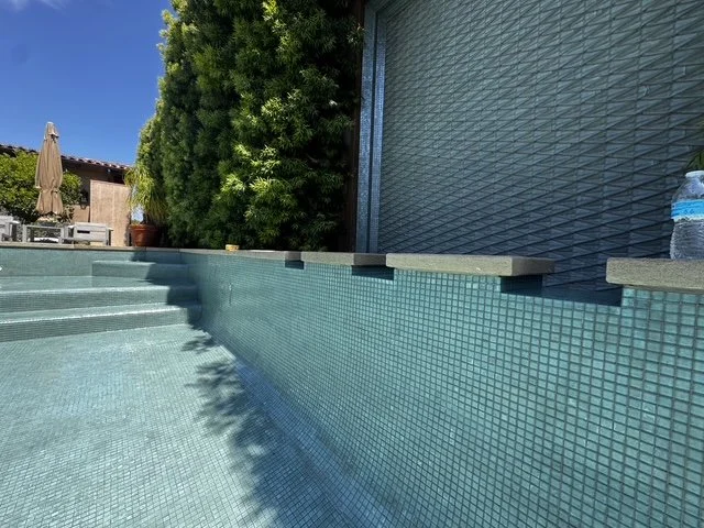Empty swimming pool with light blue mosaic tiles, surrounding steps, a large green shrub, a patio umbrella, and outdoor furniture in the background.