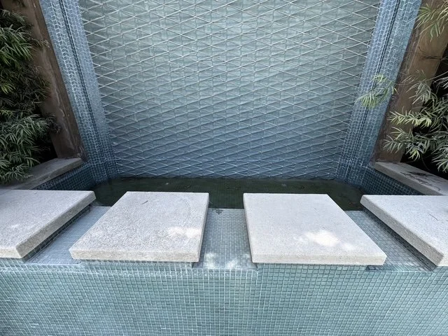 Four stone stepping tiles placed in a shallow water feature with a mosaic tile backdrop, surrounded by plants.
