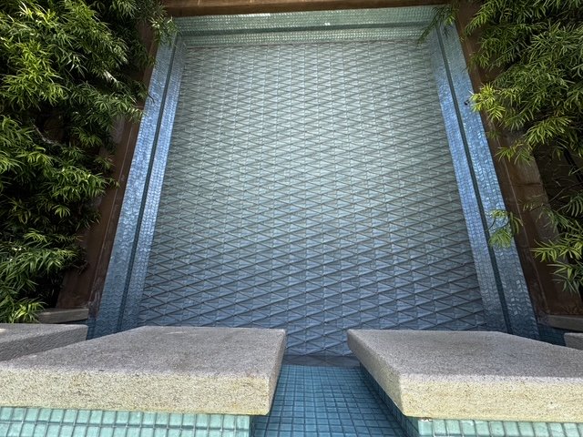 A vertical water fountain feature with water cascading down a textured surface, surrounded by lush greenery and two concrete sitting areas at the bottom.