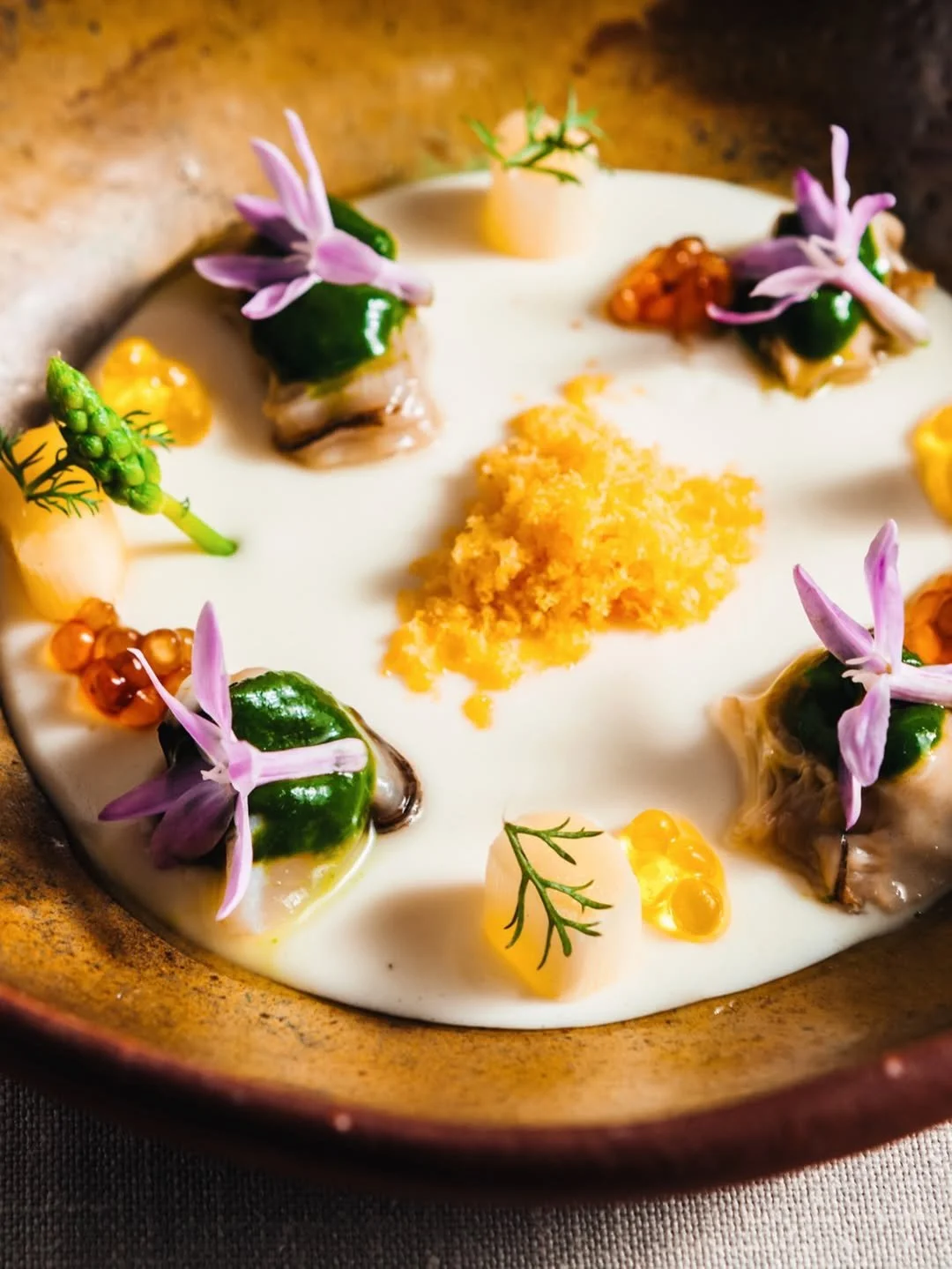A plated gourmet dish with small, wrapped portions garnished with edible flowers, herbs, a yellow crumb, and dollops of creamy sauce.