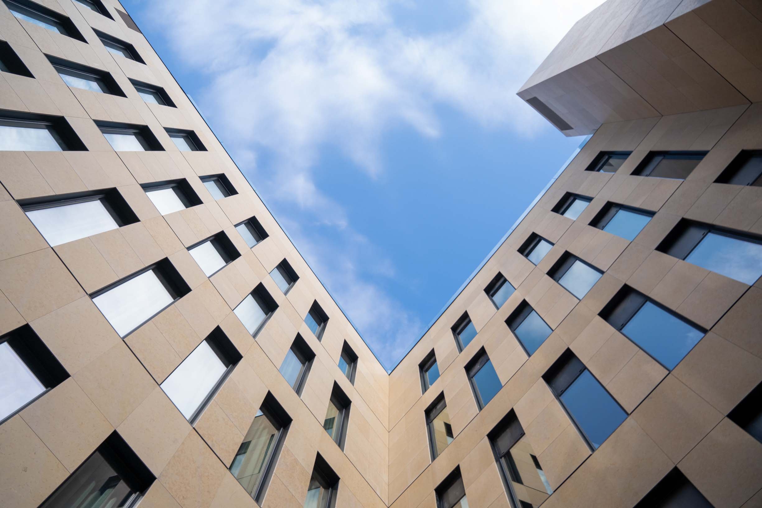 Looking up at a modern building with beige exterior and evenly spaced black-framed windows against a blue sky with scattered clouds.