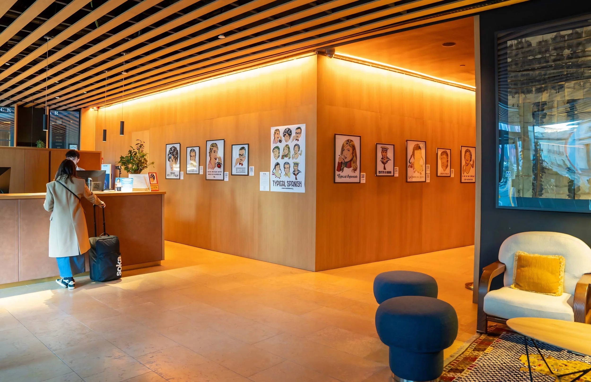 Hotel lobby with wooden wall decor, colorful posters on the wall, a reception desk with a woman checking in, seating area with modern chairs, and a large window on the right.