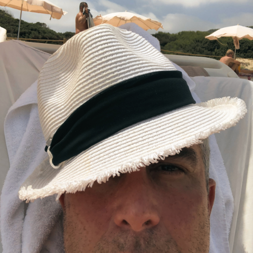 Person sunbathing on a beach with a white hat resting on their forehead, others under umbrellas in the background.