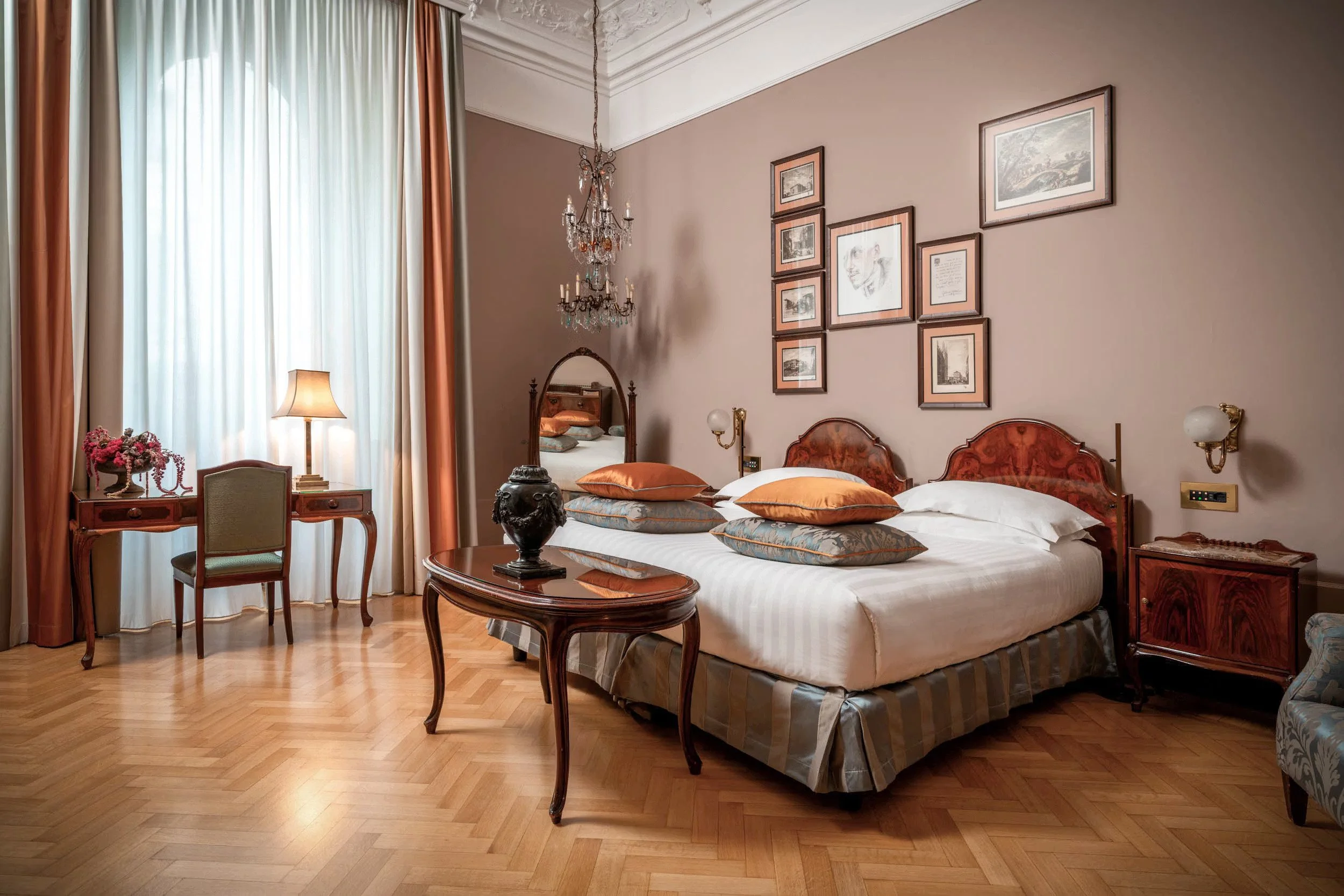 A bedroom with a wooden floor, two beds with orange and white bedding, a wooden nightstand, a mirror, framed pictures on the wall, a chandelier, a window with curtains, and a small desk with a chair and a flower arrangement.