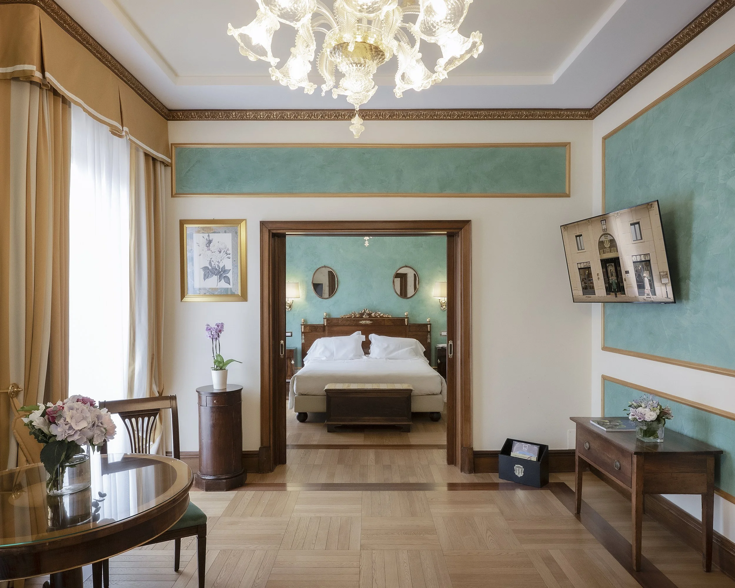 Open-concept living and bedroom area with high ceilings, elegant chandelier, large window dressed with beige curtains, green and cream-colored walls, framed artwork, wooden furniture including a coffee table with a floral arrangement, a small side table, and a flat-screen TV.
