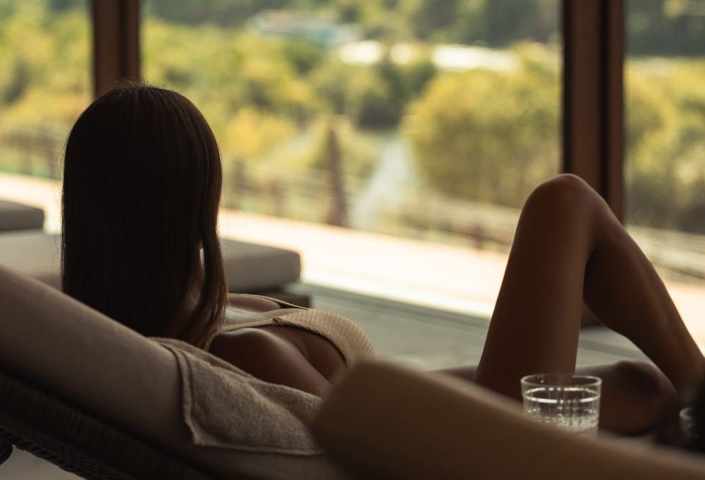 A woman with medium-length dark hair lying on a lounge chair inside, looking out a large window at a scenic view of trees and a pathway.