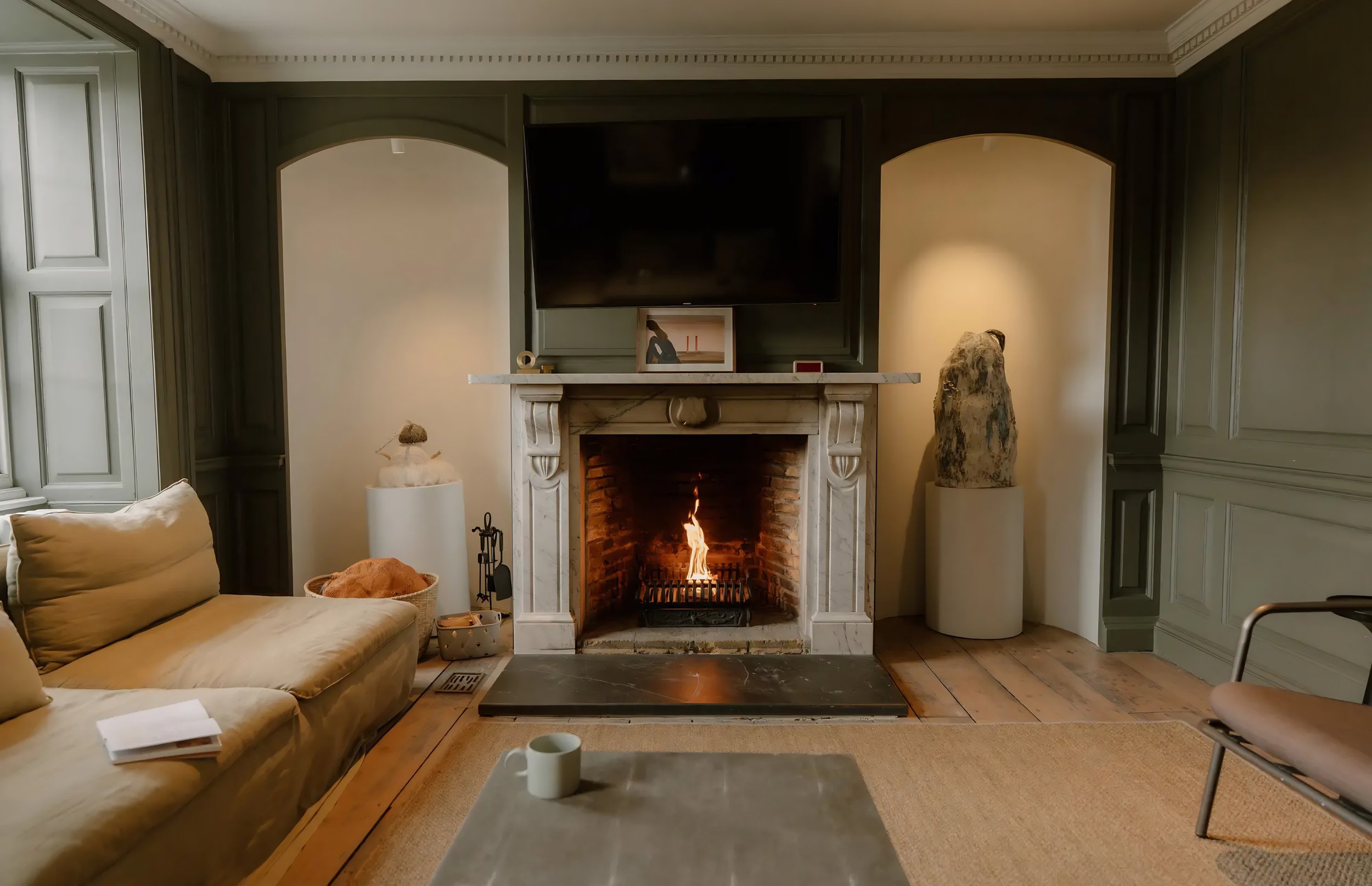 Living room with a fireplace, a beige sofa, a coffee table with a white mug, and a large flat-screen TV above the fireplace. The decor includes a tall stone sculpture on the right side of the fireplace, a small white side table with a decorative obje