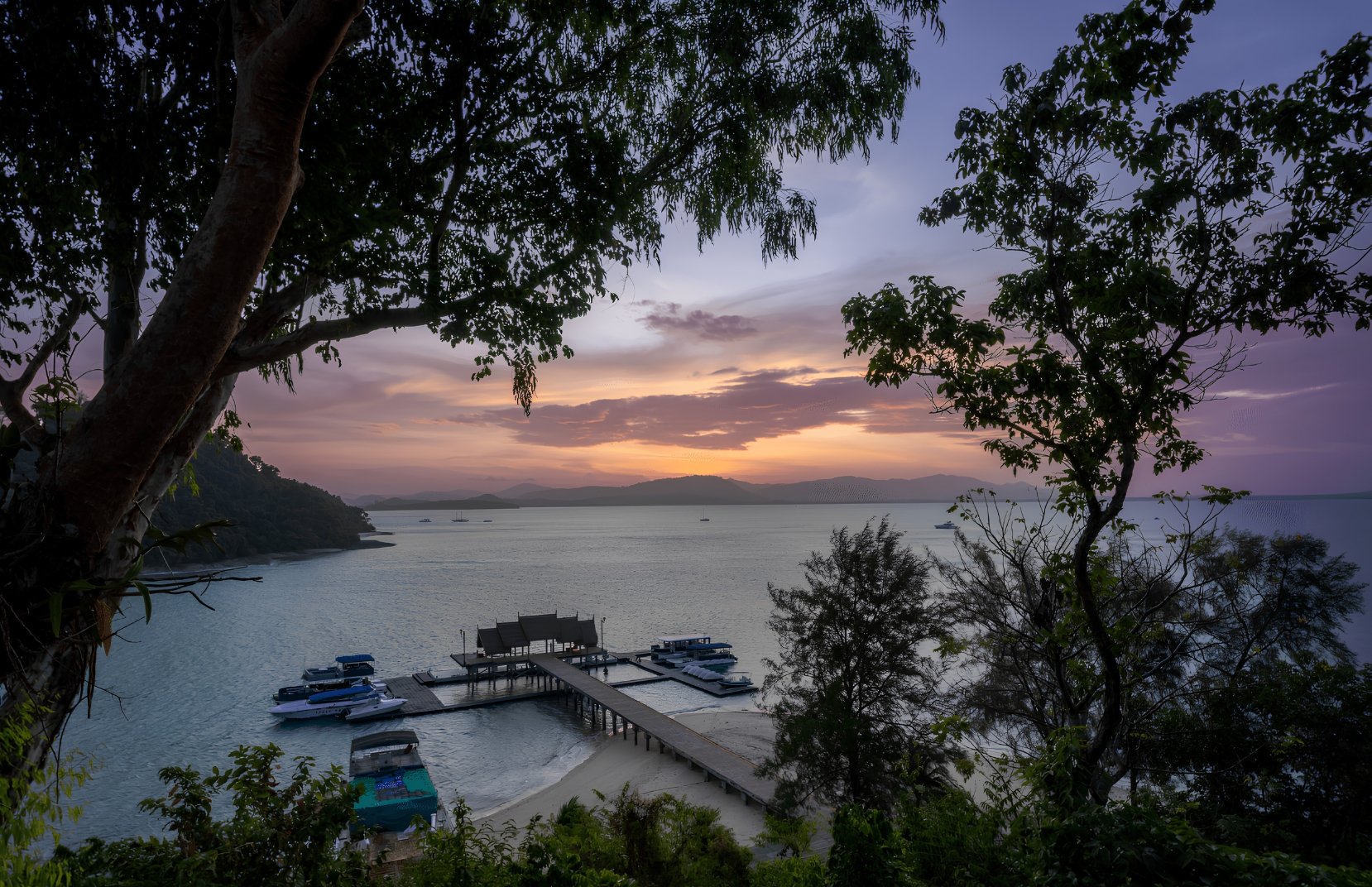View of a harbor at sunset with boats docked at a pier, trees framing the scene, and mountains in the background under a colorful sky.