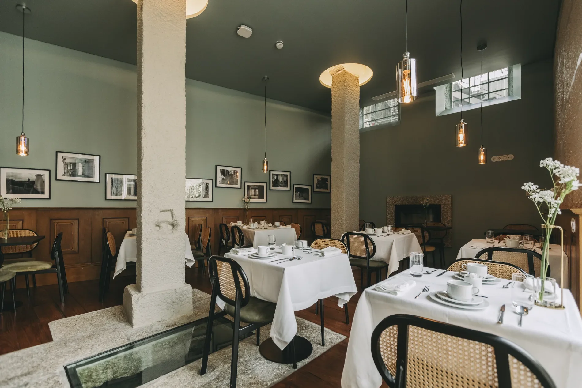 Elegant restaurant dining area with tables set for a meal, white tablecloths, and nice dishware. Hanging pendant lights and framed photos decorate the walls. Natural light filters in through high windows, creating a warm atmosphere.
