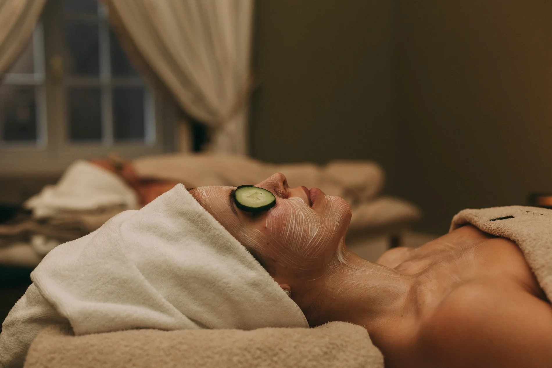 A person lying down with a face mask on, a cucumber slice on their eye, and a towel wrapped around their head, receiving a spa treatment in a cozy room.