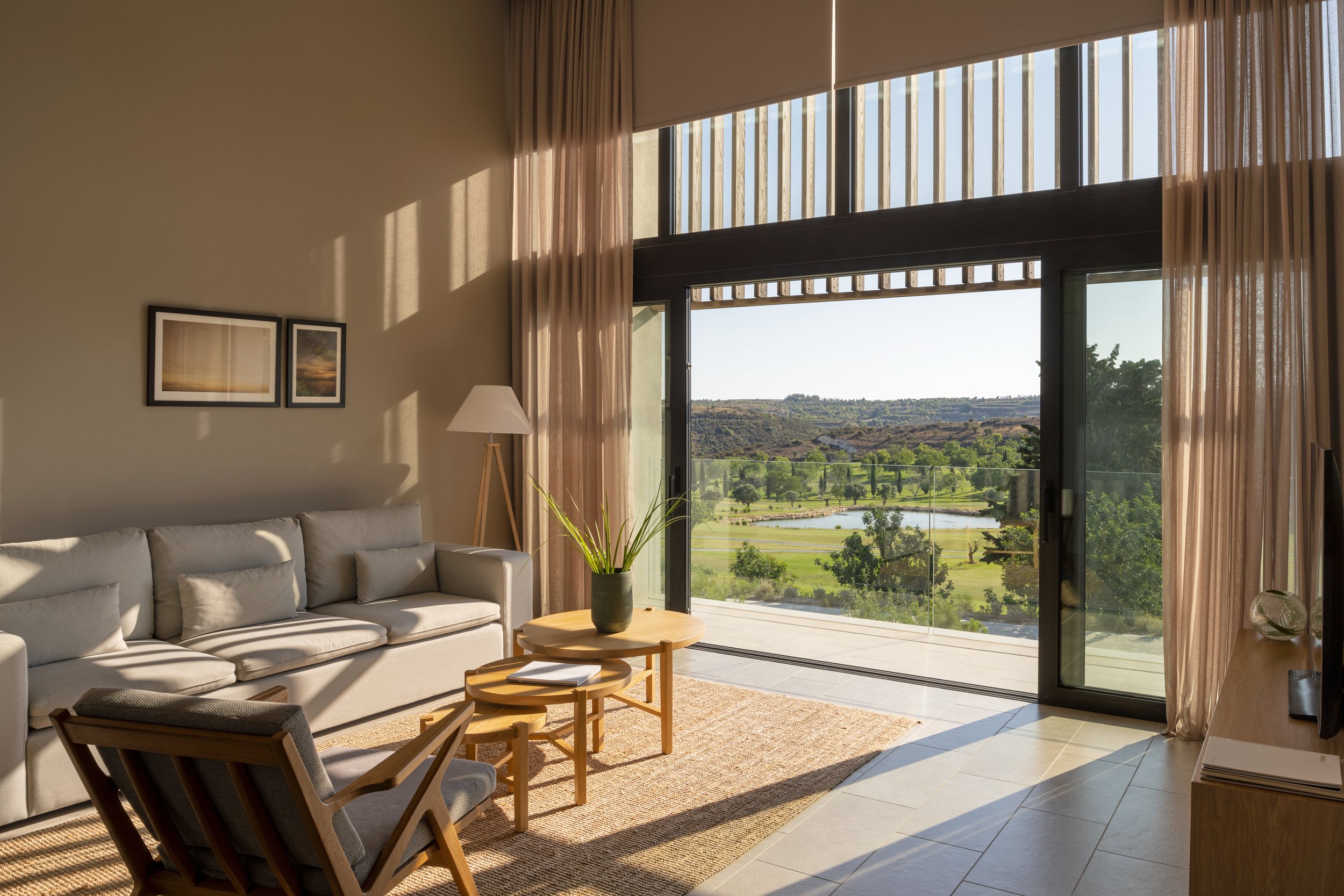 Living room with large sliding glass doors showing a view of a golf course and rolling hills, beige walls, framed artwork, a white sofa, a wooden coffee table with a potted plant, and armchairs near the table, with sunlight casting shadows.