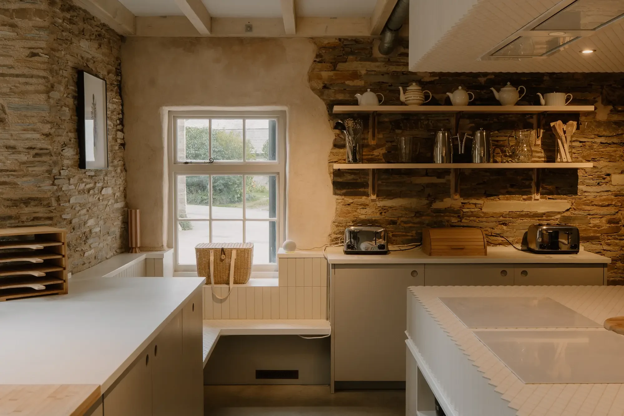 Kitchen with exposed stone and plaster brick walls, white cabinets, open wooden shelves with teapots, jars, and utensils, a window with greenery outside, and small appliances on the counter.